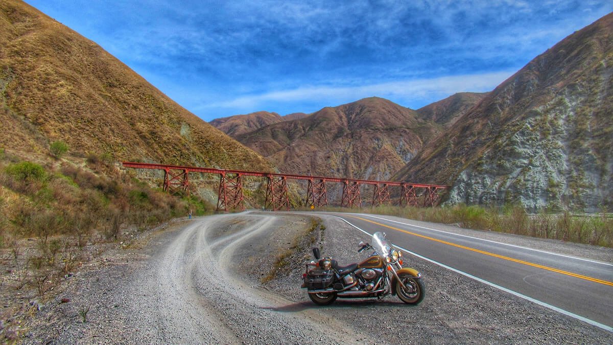 Moto Harley-Davidson chamada Formosa parada às margens da Ruta Nacional 51, com o histórico Viaducto de la Quebrada del Toro ao fundo, cercado por montanhas coloridas da Cordilheira dos Andes, em direção ao Sítio Arqueológico Tastil, no Norte da Argentina.