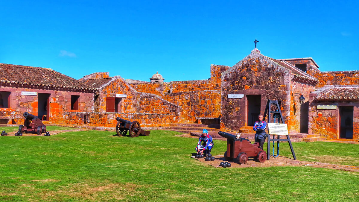 Perspectiva detalhada do Fuerte de San Miguel, Uruguai, com destaque para canhões históricos e a capela ao fundo sob um céu azul límpido.