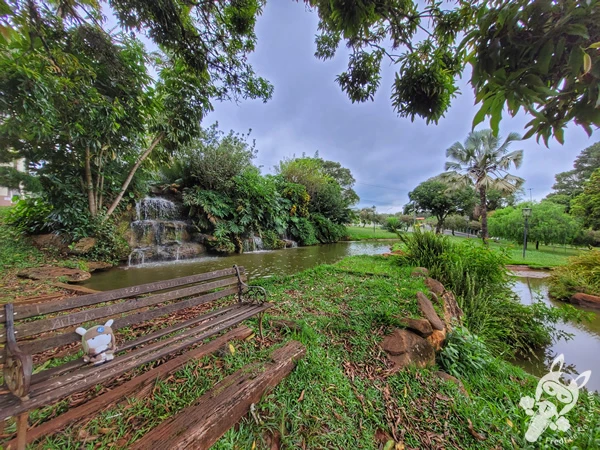 Banco sob a sombra de árvores, com uma cascata artificial ao fundo, na Praça da Cachoeira, em Holambra, São Paulo, Brasil. Foto: FredLee Na Estrada.